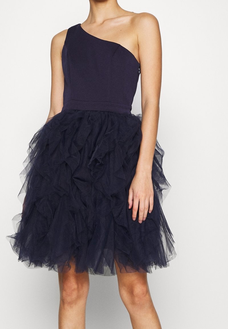 Laivastonsininen mekko, jossa on istuva, yhdellä olkapäällä oleva yläosa ja laskostettu tyllihame, jotka yhdessä luovat tekstuurisen kerrostehosteen.