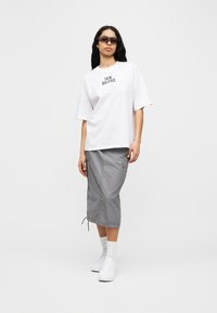 Λευκό βαμβακερό φαρδύ t-shirt με λογότυπο «New Balance», σε συνδυασμό με γκρι cargo φούστα μέχρι το γόνατο και λευκά αθλητικά παπούτσια. Φοράει σκούρα γυαλιά ηλίου.