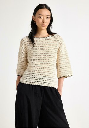 Kvinde iført en cremefarvet åbenstrikket sweater med albuelange ærmer og løse sorte bukser, stående med hænderne i lommerne foran en ensfarvet baggrund.