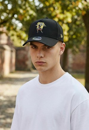 Young man wearing a black baseball cap with a colorful SpongeBob SquarePants embroidered design, standing outdoors with blurred greenery background.