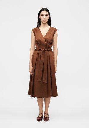 Kobieta stojąca, ubrana w brązową, bezrękawnikową sukienkę midi z dekoltem w kształcie litery V i wiązanym paskiem w talii, w połączeniu z ciemnoczerwonymi butami Mary Jane.