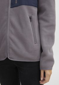 Giacca in pile grigia con un pannello superiore blu navy, caratterizzata da una tasca con zip sul lato e una finitura in tessuto testurizzato. Morbida, calda e dal taglio casual.