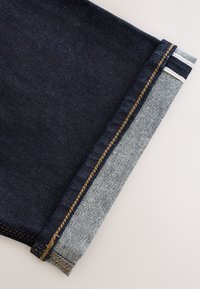 Risvolto della gamba dei pantaloni in denim blu scuro piegato per mostrare il tessuto a righe bianche e nere all'interno con cuciture arancioni e gialle.