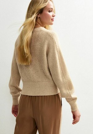 Suéter beige de punto con dobladillo acanalado, corte relajado y mangas largas, combinado con pantalones marrones. Textura suave y diseño informal.