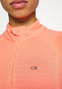 Pull-over à manches longues corail vif avec col zippé, tissu texturé et logo "ck" brodé en noir sur le côté gauche. Coutures accentuées visibles.