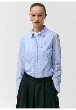 Kobieta ubrana w jasnoniebieską koszulę na guziki z drobnymi ciemnymi kwiatowymi wzorami i czarną plisowaną spódnicę, stojąca na tle jednolitego białego tła.