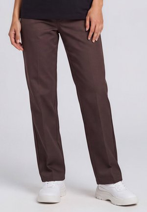 Personne portant un pantalon marron foncé droit et des baskets blanches épaisses, debout devant un fond clair uni.