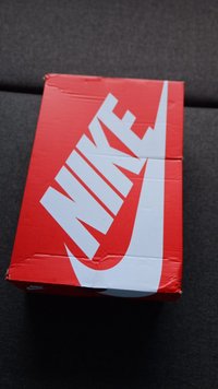 Nike'i kingituskarp erksas punases värvuses, millel on suur valge swoosh-logo ja tekst "NIKE". Karp näitab kulumise märke, sealhulgas kortse ja kriimustusi.