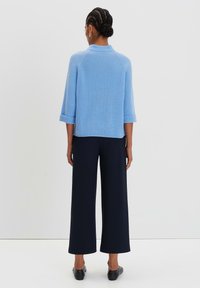 Hellblaues, geripptes Strickoberteil mit Dreiviertelärmeln, kombiniert mit weit geschnittenen dunkelblauen Hosen, vor einem schlichten Hintergrund stehend.