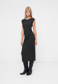 Čierne bezrukávové šaty s bielymi bodkami, po kolená, priliehavý strih. Outfit doplňujú čierne čižmy po kolená.