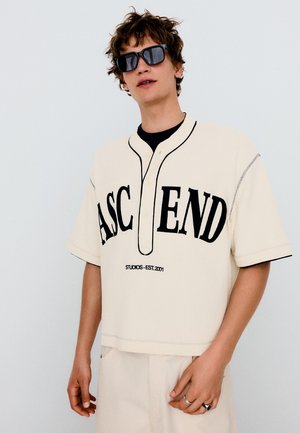 Nuori henkilö käyttää ylisuurta kermaista baseball-tyylistä paitaa, jossa lukee "ASCEND" ja mustia aurinkolaseja yksiväristä taustaa vasten.