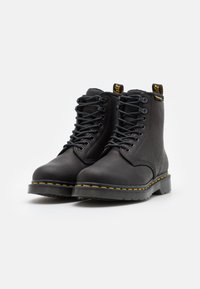 Botas de cuero negras hasta el tobillo con costuras amarillas, punta redonda y cordones. Presenta tiradores en bucle en el talón para facilitar su puesta.