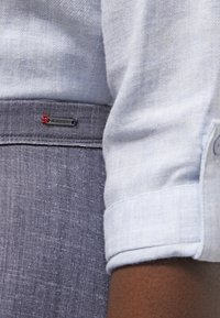 Camisa de manga larga azul clara con tejido sutilmente texturizado, combinada con pantalones grises que tienen una etiqueta de logo roja y patrones de tela en contraste.