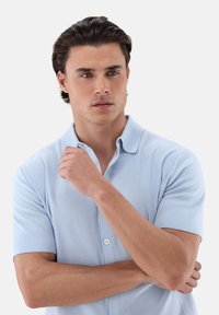 Polo blu chiaro realizzato in cotone liscio, con colletto a bottoni, maniche corte e una vestibilità comoda. Braccia incrociate davanti.