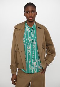 Brown short jacket with a collar, layered over a green striped shirt featuring white flowers and dark buttons, paired with matching bottoms.