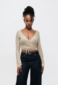 Béžs wrap tops ar garām piedurknēm un dziļu V-veida kaklu, kombinēts ar tumšām platām džinsa bikšu kājām un augstu jostasvietu. Fakturēts audums ar sasiešanas detaļu viduklī.