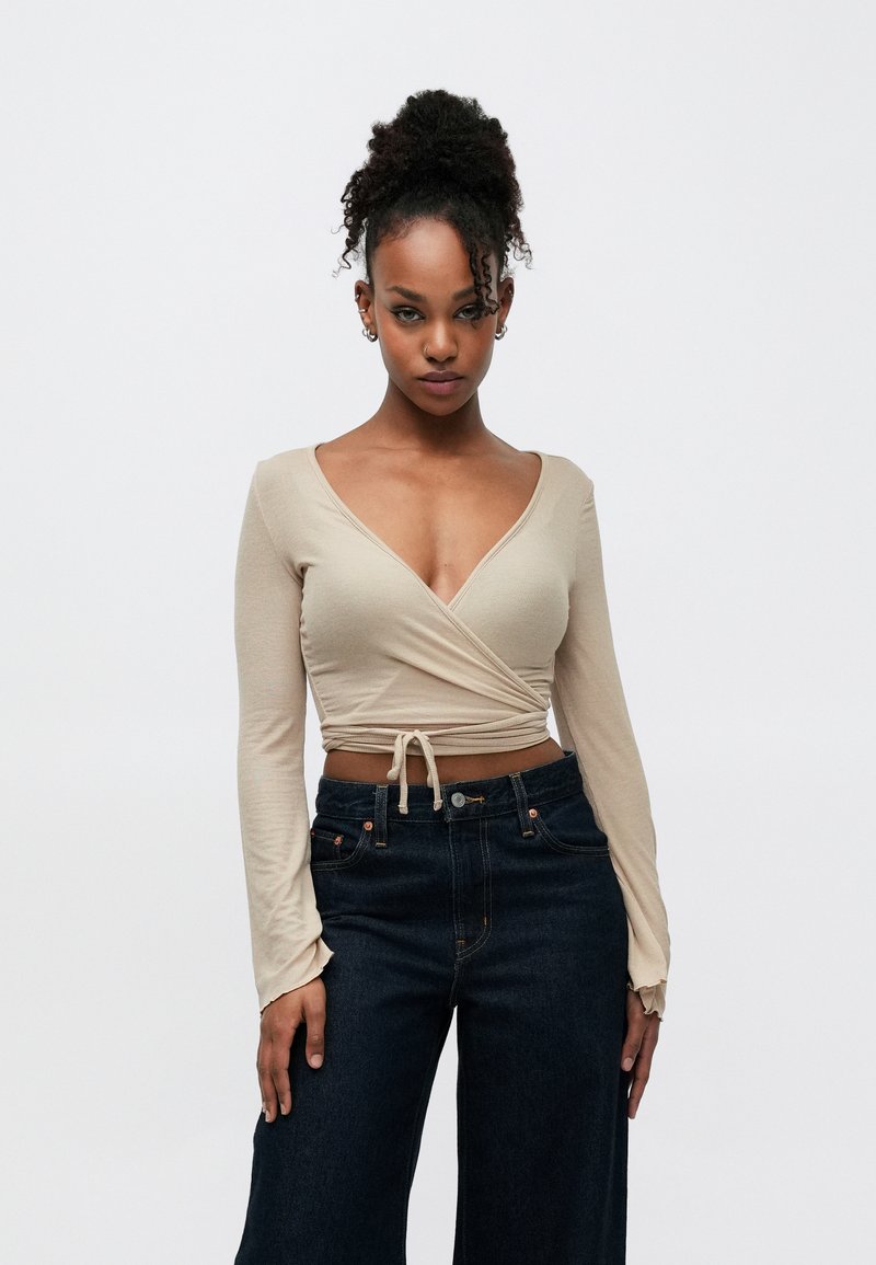 Béžs wrap tops ar garām piedurknēm un dziļu V-veida kaklu, kombinēts ar tumšām platām džinsa bikšu kājām un augstu jostasvietu. Fakturēts audums ar sasiešanas detaļu viduklī.