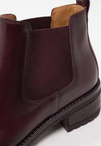 Burgundin värinen nahkasaapas nilkkakorkeudella, jossa on elastiset sivupaneelit, teksturoitu ruskea pohja ja erottuva ompelu. Pyöristetty kärki ja keskikorkea korko.