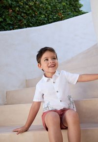 Chemise blanche à manches courtes avec broderie de palmier et boutons marron, associée à un short rose. Assis sur des marches courbes beige.