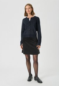 Cardigan tricotat bleumarin cu guler rotund și închidere cu nasturi, asortat cu o fustă neagră din catifea. Modelul poartă ciorapi negri și mocasini.