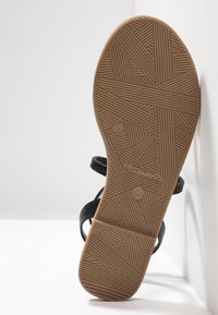 Musta sandaali, jossa on teksturoidun kumipohjan geometrinen kuvio ja minimalistinen muotoilu. Remmit ovat pehmeää materiaalia mukavuuden takaamiseksi.