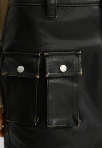 Svart läderkjol med två framfickor med tryckknappar, synlig söm och en slät yta med lätt glans.