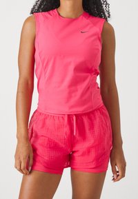 Top deportivo sin mangas de color rosa con cuello redondo y logotipo, combinado con pantalones cortos rosas que presentan un patrón de cuadrícula texturizada y una cinturilla elástica.