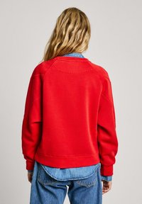 Felpa rossa con una vestibilità morbida, scollatura rotonda e maniche raglan. Indossata sopra una camicia di jeans azzurra. Texture liscia, design minimale.