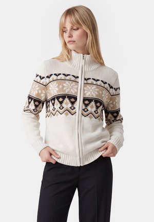 Junge Frau trägt einen cremefarbenen Pullover mit Reißverschluss mit schwarzen und beigefarbenen geometrischen und Schneeflockenmuster, kombiniert mit schwarzen Hosen, vor einem schlichten Hintergrund stehend.
