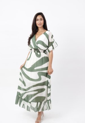 Woman with long dark hair wearing a white and green patterned maxi dress with ruffle hem and clear strap heels, posing against a white background.