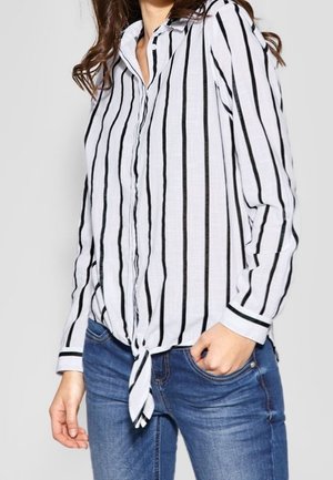 Vrouw die een wit shirt met lange mouwen en zwarte verticale strepen draagt, geknoopt in de taille, gecombineerd met blauwe spijkerbroek.