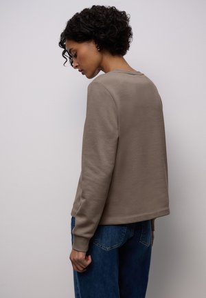 Jasnobrązowa bluza o luźnym kroju, wykonana z fakturowanego materiału, z okrągłym dekoltem i prążkowanymi mankietami, zestawiona z niebieskimi jeansami o szerokich nogawkach.