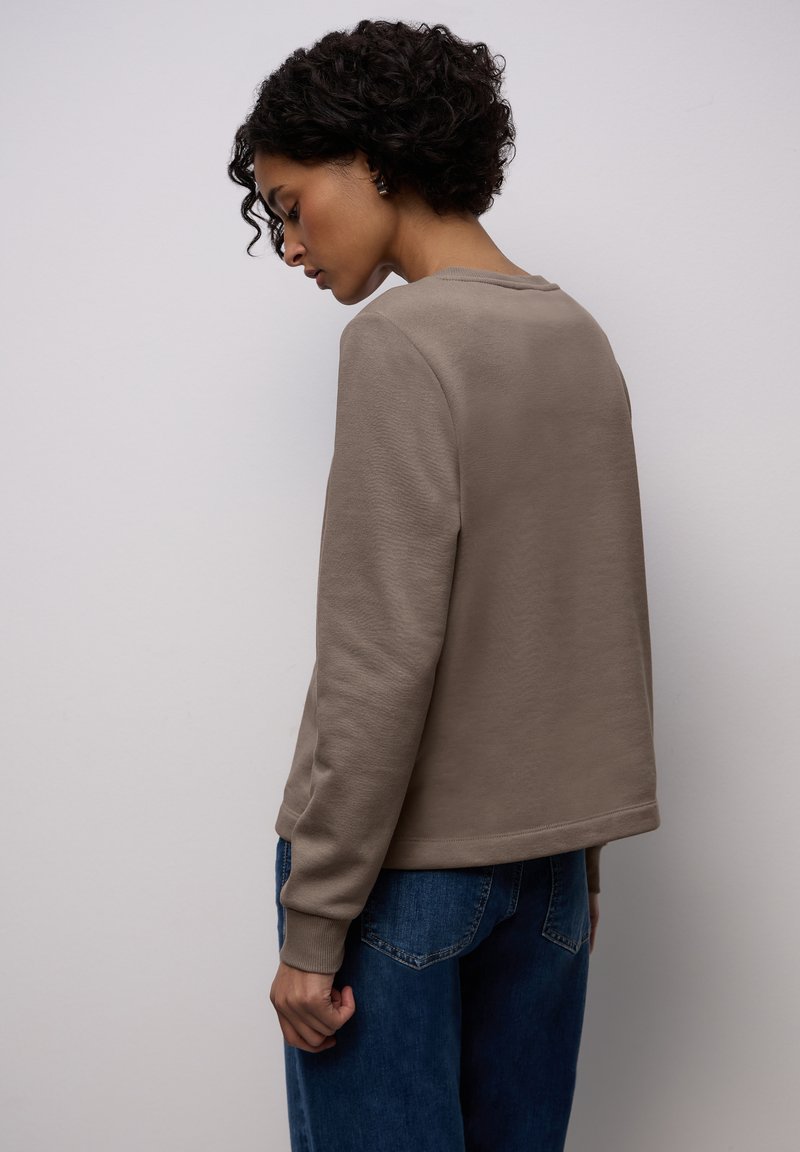 Jasnobrązowa bluza o luźnym kroju, wykonana z fakturowanego materiału, z okrągłym dekoltem i prążkowanymi mankietami, zestawiona z niebieskimi jeansami o szerokich nogawkach.