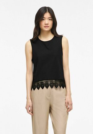 Femme aux cheveux lisses et foncés portant un haut noir sans manches avec un ourlet en dentelle et un pantalon beige à jambes larges, debout sur un fond blanc.