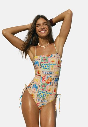 Lächelnde Frau mit langen Haaren, die einen bunten Einteiler mit Frucht- und Palmenmuster trägt und mit erhobenen Armen hinter dem Kopf posiert.