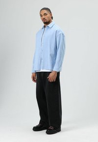 Lichtblauw overhemd van zachte stof gecombineerd met losse zwarte broek en zwarte schoenen met roze veters. Model staat tegen een neutrale achtergrond.