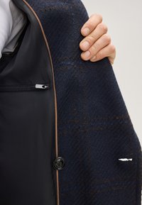 Marineblaue Wolljacke mit Fischgrätstruktur, kontrastierendem schwarzem Futter, braunen Zierleisten und zwei Reißverschlusstaschen; auffällige kleine Rissstelle im Stoff.