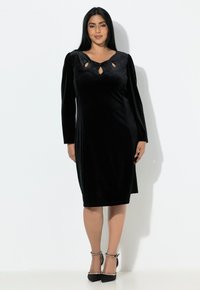 Czarna aksamitna sukienka o dopasowanej sylwetce, z długimi bufiastymi rękawami i wycięciami w kształcie klucza przy dekolcie. Materiał ma delikatny połysk.