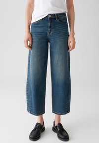 Persoon in cropped wide-leg blauwe jeans, wit topje en zwarte veterschoenen tegen een effen lichte achtergrond.