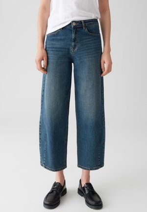 Personne portant un jean large coupe courte, un haut blanc et des chaussures à lacets noires sur un fond clair.