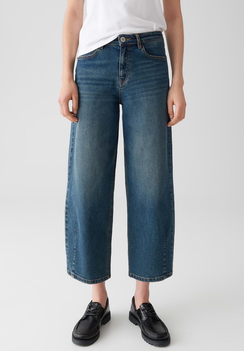 Persoon in cropped wide-leg blauwe jeans, wit topje en zwarte veterschoenen tegen een effen lichte achtergrond.