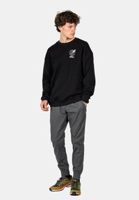 Sudadera negra de cuello redondo con logo blanco de globo, combinada con pantalones gris con puños elásticos y zapatillas deportivas verdes con acentos naranjas.