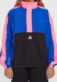 Personne portant un pull polaire bicolore bleu, rose et noir avec une fermeture éclair quart et le logo O'Neill sur la poitrine.