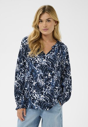 Tmavomodrá blúzka s kvetinovým vzorom v bielej a svetlejšej modrej, s výstrihom do V a dlhými rukávmi s nariasenými manžetami.