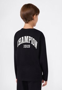 Dziecko z krótkimi brązowymi włosami, odwrócone plecami, ubrani w czarną bluzę z długim rękawem z napisem "CHAMPION 1919" w białym kolorze na plecach.