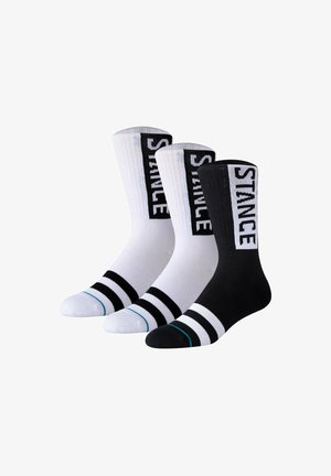 Drei knielange Socken, zwei weiße und eine schwarze, jeweils mit fettgedrucktem "STANCE" Schriftzug an der Seite und zwei schwarzen oder weißen Streifen nahe den Zehen.