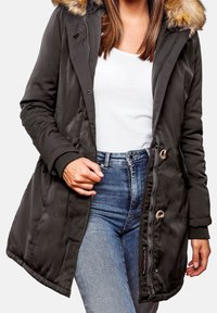 Parka verde con capucha forrada de piel, cierre con cremallera y botones, puños de canalé y bolsillos laterales; usado sobre una camisa blanca y unos jeans de mezclilla.