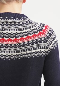 Person som bär en marinblå tröja med ett stickat Fair Isle-mönster i vitt och rött över den övre delen av ryggen och axlarna.