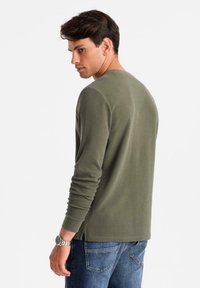 Mann trägt ein olivgrünes Langarmhemd und eine blaue Jeans, leicht zur Seite geneigt, mit einer silbernen Armbanduhr am linken Handgelenk.