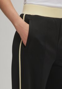 Schwarze Hose mit breitem beigen Bund, seitlichem Taschen und einem kontrastierenden beigen Streifen entlang des Beins. Glatte Textur.